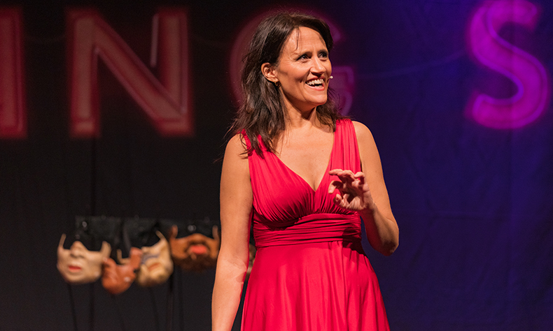 A woman with dark brown hair in a red dress smiles as she looks at an off-camera audience. Behind her is a set of masks each with the lower half of a different face on. Promotional image from Nina Conti The Dating Show.