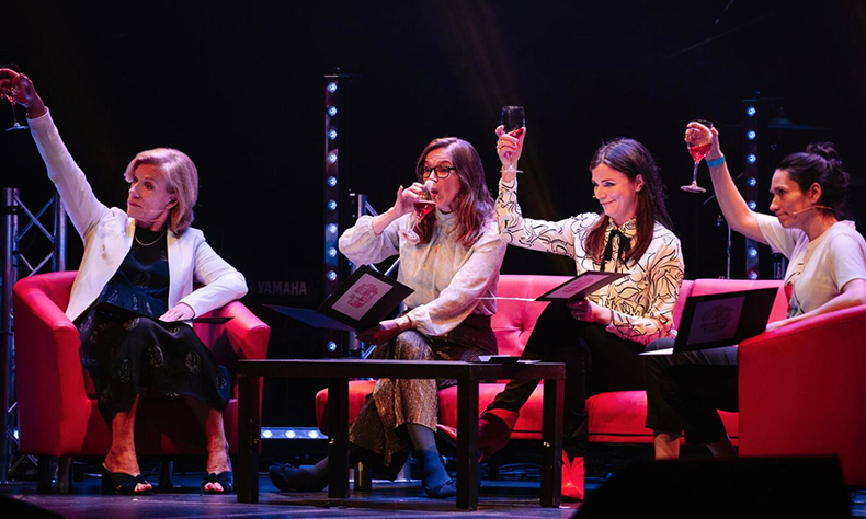 Performance photo of The Guilty Feminist. Four light-skinned women sat on sofas with a small table in front of them hold partially-full wine glasses and gesture off-camera to the audience.