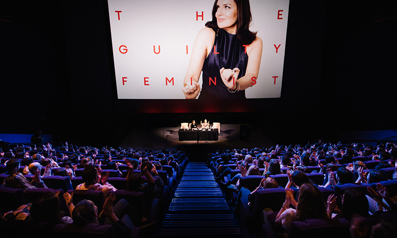 Large audience watching The Guilty Feminist. They are watching two women sat at a table in the distance underneath a large projected image of a white woman in a blue dress with brown hair mid-dance with the words The Guilty Feminist overlayed.
