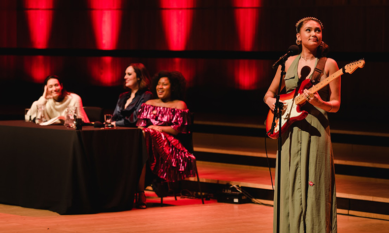 Performance photo from The Guilty Feminist. At a table, two light-skinned women and one dark-skinned woman are laughing. One of the women has her head resting in her palm and is watching a woman in a green dress and a headband play the electric guitar.