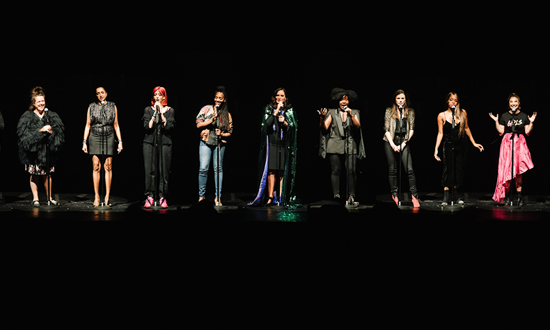Nine female performers stood on a stage with microphones in front of them. They are each illuminated by a spotlight and otherwise stood in darkness.