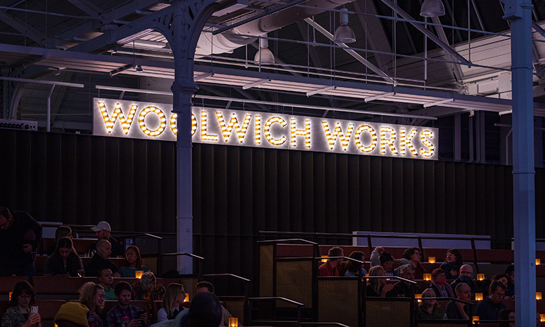 venue banner at woolwich works reading the name of the venue. Audience members lit by table candles sat in front.